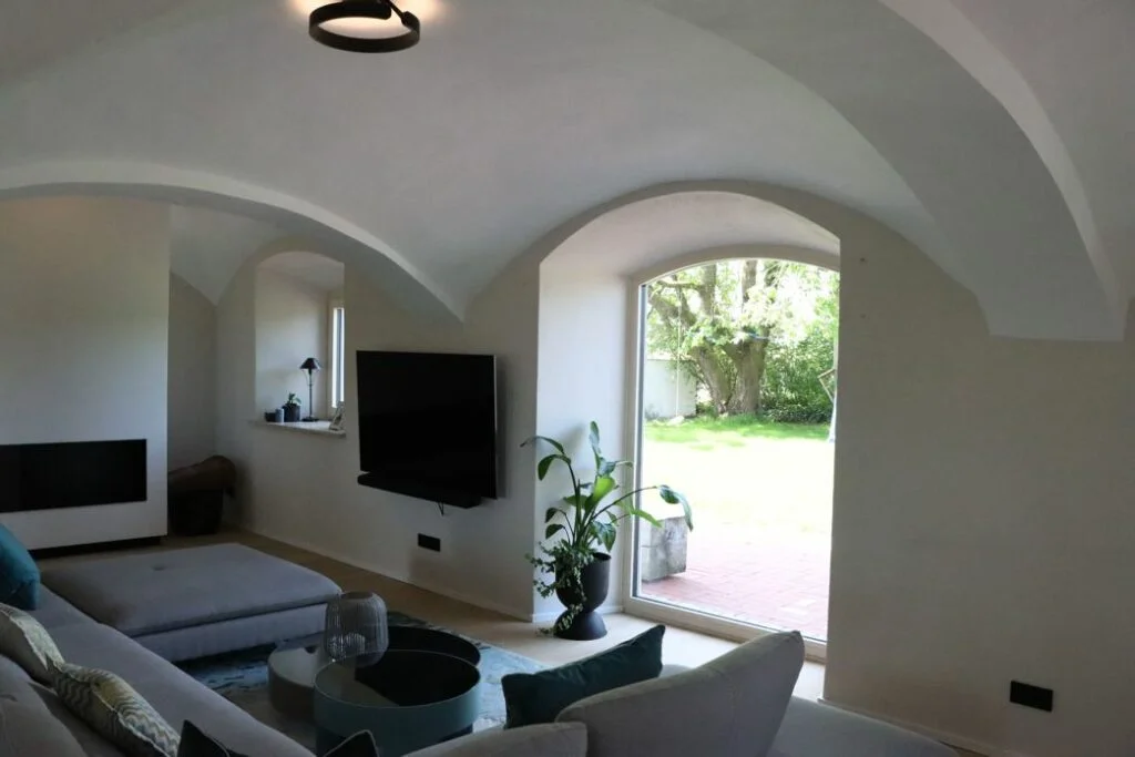 Ein helles Wohnzimmer mit gewölbten Decken, einem an der Wand montierten Fernseher, einer großen Fenstertür, die in einen Garten führt, einem grauen Sektionssofa, Zimmerpflanzen und neutralen Wänden - das sind Referenzen Bauelemente in der modernen Innenarchitektur.