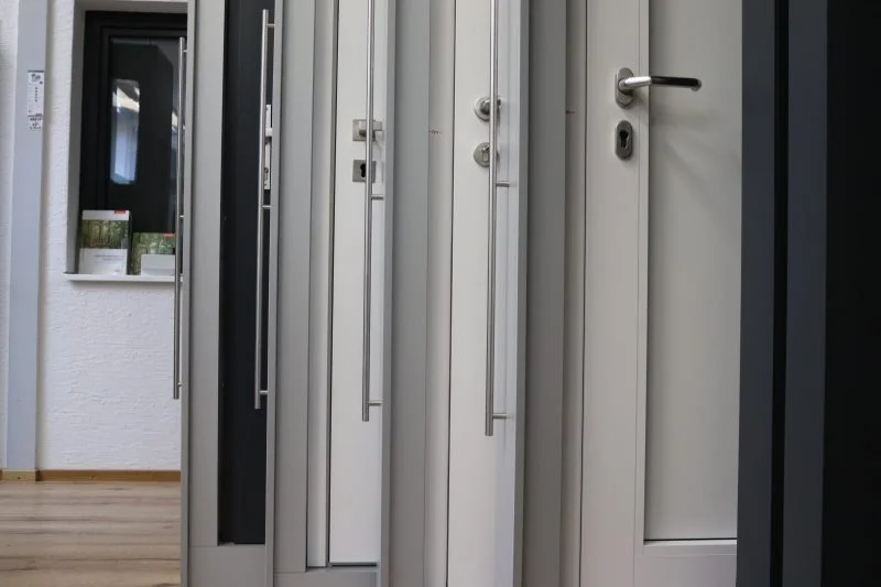 Im Ausstellungsraum der Bauelemente Ausstellung sind mehrere moderne Innentüren, meist in Weiß und Grau, aufrecht stehend ausgestellt. Im Hintergrund links ist ein kleines Fenster mit Topfpflanzen auf der Brüstung zu sehen.